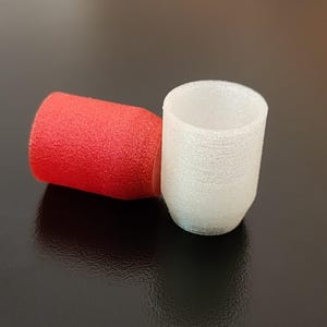 May include: A red cylindrical object and a white cylindrical object are positioned on a dark surface. The red object is textured, while the white object has a smooth, slightly textured appearance. The objects appear to be small caps or stoppers.