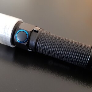 Könnte beinhalten: Schwarze Taschenlampe mit strukturierter Griff und blauem Knopf. Die Taschenlampe hat eine weiße Abdeckung über dem Lichtende. Die Taschenlampe liegt auf einer dunklen Oberfläche.