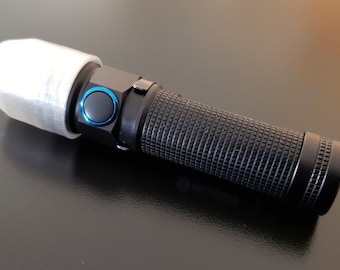 以下が含まれることがあります： 黒色の懐中電灯。グリップはテクスチャ加工され、青いボタンが付いています。懐中電灯の光の端には白いカバーが付いています。懐中電灯は暗い表面にあります。