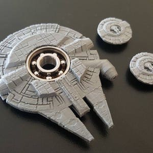 May include: A gray 3D printed fidget spinner shaped like the Millennium Falcon from Star Wars. The spinner has a silver bearing in the center and three smaller gray spinners on the sides.