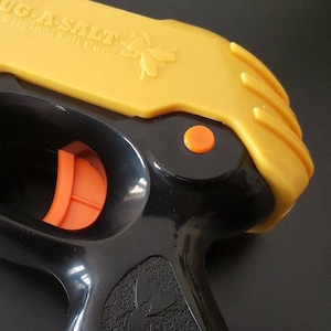 May include: A yellow and black plastic toy gun with the words "Bug-A-Salt The Original Salt Gun" printed on the top. The gun has an orange trigger and a black barrel.
