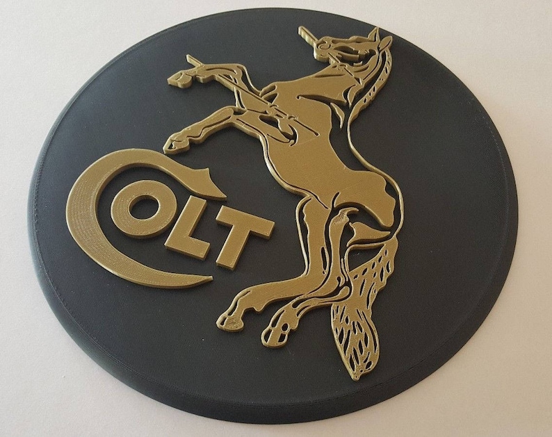 May include: A black circular plaque with a gold-coloured Colt logo and a stylised horse design. The logo reads "COLT" in all caps.