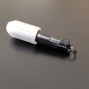 May include: A black Olight i3E EOS keychain flashlight with a white protective sleeve. The flashlight has a silver keyring attached.