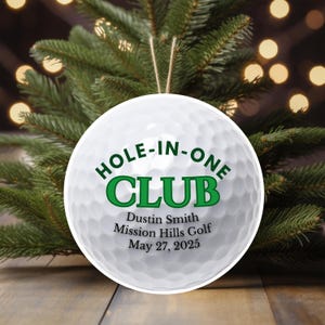 May include: White golf ball ornament with the text "HOLE-IN-ONE CLUB" in green. The ornament includes the name "Dustin Smith", "Mission Hills Golf", and the date "May 27, 2025". The ornament hangs from a string, with a blurred background of a Christmas tree.