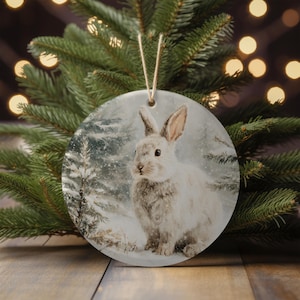 Puede incluir: Adorno navideño redondo de cerámica con una escena invernal con un conejo blanco en un bosque nevado. El adorno tiene una cuerda para colgar y se exhibe frente a un árbol de Navidad.