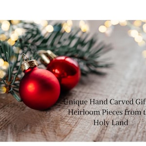 May include: Two smooth, round, red Christmas ornaments with gold-colored caps resting near green pine needles. The text "Unique Hand Carved Gifts & Heirloom Pieces from the Holy Land" is printed in black on the lower right side of the image.