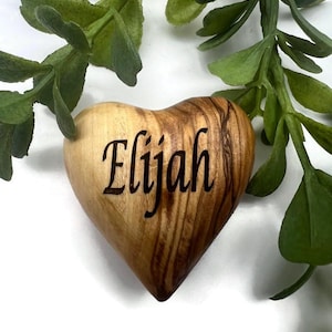 May include: A smooth, heart-shaped wooden object with a natural light and dark brown grain pattern. The name Elijah is engraved in a dark, cursive script across the center of the heart. The object is positioned against a white background.