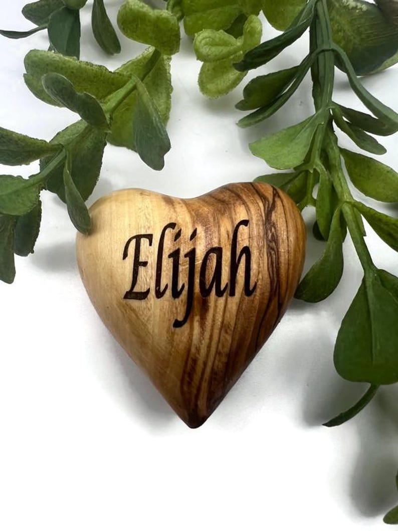 May include: A smooth, heart-shaped wooden object with a natural light and dark brown grain pattern. The name Elijah is engraved in a dark, cursive script across the center of the heart. The object is positioned against a white background.
