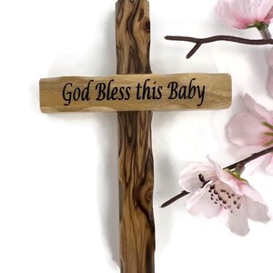 May include: Wooden cross with the inscription "God Bless this Baby". The cross is made of light brown wood with dark brown grain patterns. Pink flowers are in the background.