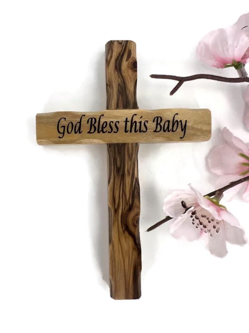 May include: Wooden cross with the inscription "God Bless this Baby". The cross is made of light brown wood with dark brown grain patterns. Pink flowers are in the background.