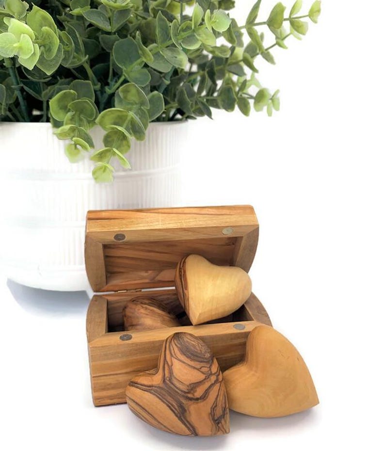 May include: A small, rectangular wooden chest with a hinged lid, containing three smooth, heart-shaped wooden objects. The hearts display natural wood grain patterns in shades of light and dark brown. The chest is crafted from light-toned wood.