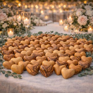 May include: A large collection of small, smooth, hand-carved wooden hearts made from olive wood, featuring distinct brown grain patterns. The hearts are arranged on a light-colored fabric surface, interspersed with green eucalyptus leaves.