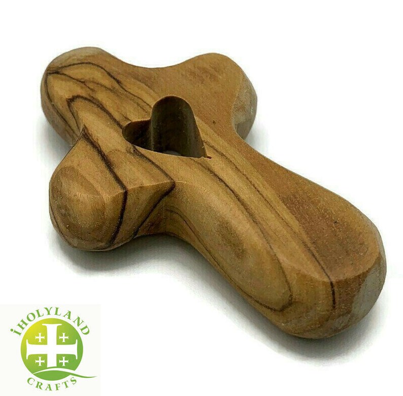 May include: A small, wooden cross with a heart-shaped cutout in the center. The cross is made of olive wood and has a natural, brown color.