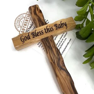 May include: A wooden cross with the inscription "God Bless this Baby" in black lettering. The cross is made of light brown wood with dark brown grain patterns. The background includes greenery and a white surface.