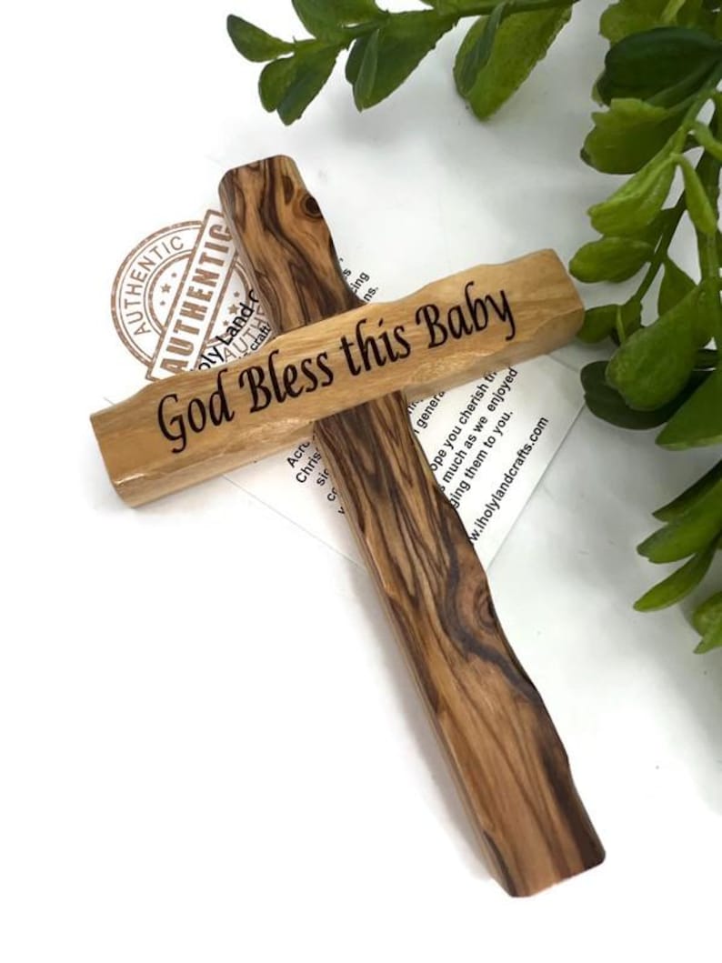 May include: A wooden cross with the inscription "God Bless this Baby" in black lettering. The cross is made of light brown wood with dark brown grain patterns. The background includes greenery and a white surface.