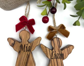 May include: Two wooden angel ornaments, each with a name inscribed: "Mia" and "Liam". They hang by twine, one with a red velvet bow, the other with a tan bow. The background features greenery and red berries. The angels have outstretched wings.