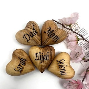 May include: Five smooth, heart-shaped wooden objects with natural grain patterns, each engraved with a name or title: Dad, Mom, Elijah, Sarah, and Selena. The hearts are arranged in a cluster, accompanied by a small branch with light pink flowers.