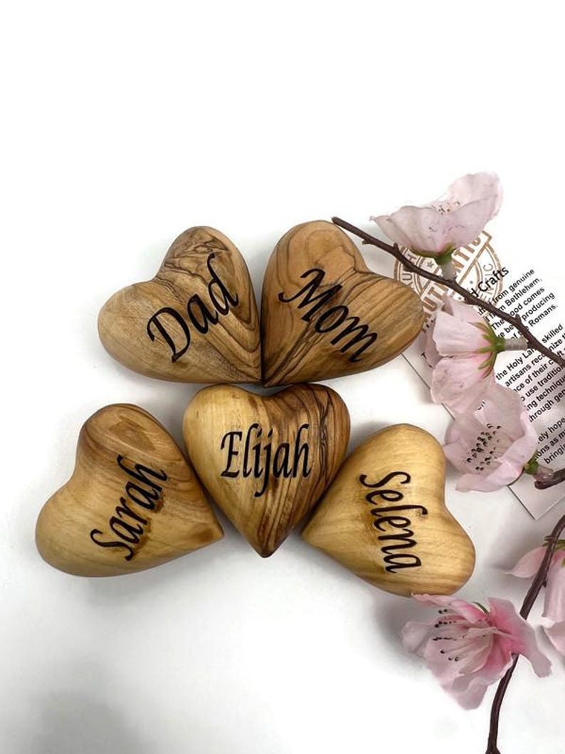 May include: Five smooth, heart-shaped wooden objects with natural grain patterns, each engraved with a name or title: Dad, Mom, Elijah, Sarah, and Selena. The hearts are arranged in a cluster, accompanied by a small branch with light pink flowers.
