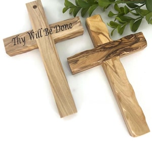 May include: Two wooden crosses with a natural wood grain pattern. One cross has the phrase "Thy Will Be Done" engraved on it. The crosses are a light brown color and are set against a white background.