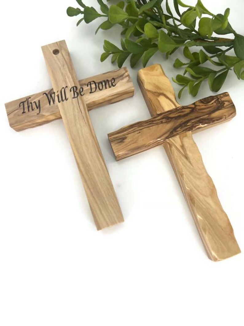 May include: Two wooden crosses with a natural wood grain pattern. One cross has the phrase "Thy Will Be Done" engraved on it. The crosses are a light brown color and are set against a white background.