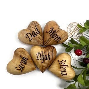 May include: Five smooth, heart-shaped wooden objects with natural grain patterns, each engraved with a name or title. The hearts are labeled Dad, Mom, Sarah, Elijah, and Selena. A small tag with text and green foliage with red berries are visible.