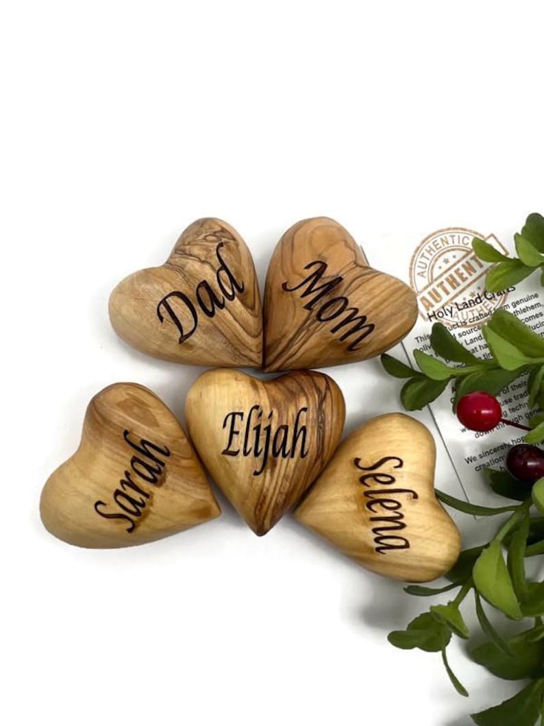 May include: Five smooth, heart-shaped wooden objects with natural grain patterns, each engraved with a name or title. The hearts are labeled Dad, Mom, Sarah, Elijah, and Selena. A small tag with text and green foliage with red berries are visible.