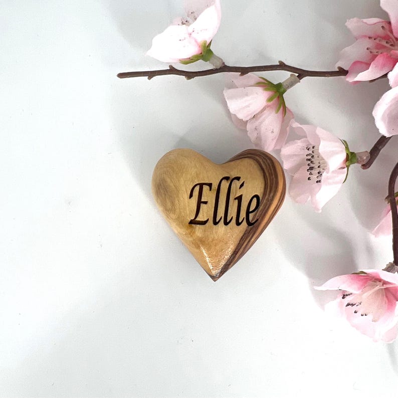 May include: A smooth, heart-shaped wooden object with a natural grain pattern, featuring the name Ellie engraved in a dark, elegant script font, positioned next to a delicate branch with light pink cherry blossom flowers.