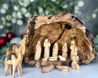May include: A nativity scene set in a carved olive wood tree trunk with a manger, baby Jesus, Mary, Joseph, three wise men, a shepherd, two camels, and a sheep.