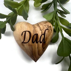 May include: A heart-shaped wooden object with a smooth, polished finish and visible natural grain patterns. The word "Dad" is engraved in a dark, elegant script font across the center of the heart, which is positioned against a white background.