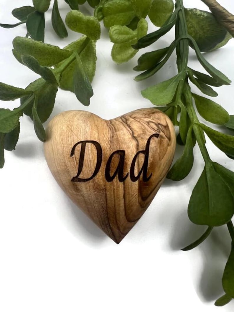 May include: A heart-shaped wooden object with a smooth, polished finish and visible natural grain patterns. The word "Dad" is engraved in a dark, elegant script font across the center of the heart, which is positioned against a white background.