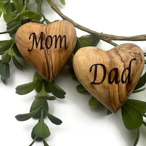 May include: Two smooth, heart-shaped wooden objects with natural grain patterns. One heart is engraved with the word Mom in a dark script font, and the other heart is engraved with the word Dad in the same dark script font, positioned near green leaves.
