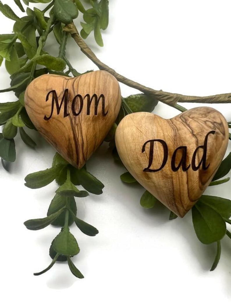 May include: Two smooth, heart-shaped wooden objects with natural grain patterns. One heart is engraved with the word Mom in a dark script font, and the other heart is engraved with the word Dad in the same dark script font, positioned near green leaves.