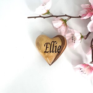 May include: A smooth, polished wooden heart ornament featuring the name Ellie engraved in a dark, elegant script font. The heart displays natural light and dark wood grain patterns and is positioned next to delicate, pale pink cherry blossom flowers.