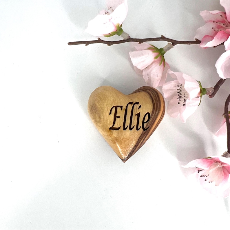 May include: A smooth, polished wooden heart ornament featuring the name Ellie engraved in a dark, elegant script font. The heart displays natural light and dark wood grain patterns and is positioned next to delicate, pale pink cherry blossom flowers.