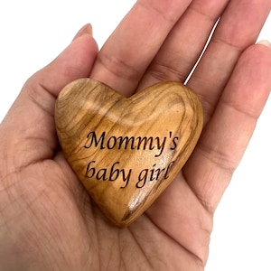 May include: A smooth, polished heart-shaped object carved from light brown wood with a visible natural grain pattern. The text "Mommy's baby girl" is engraved in a dark, cursive font across the center of the wooden heart surface.