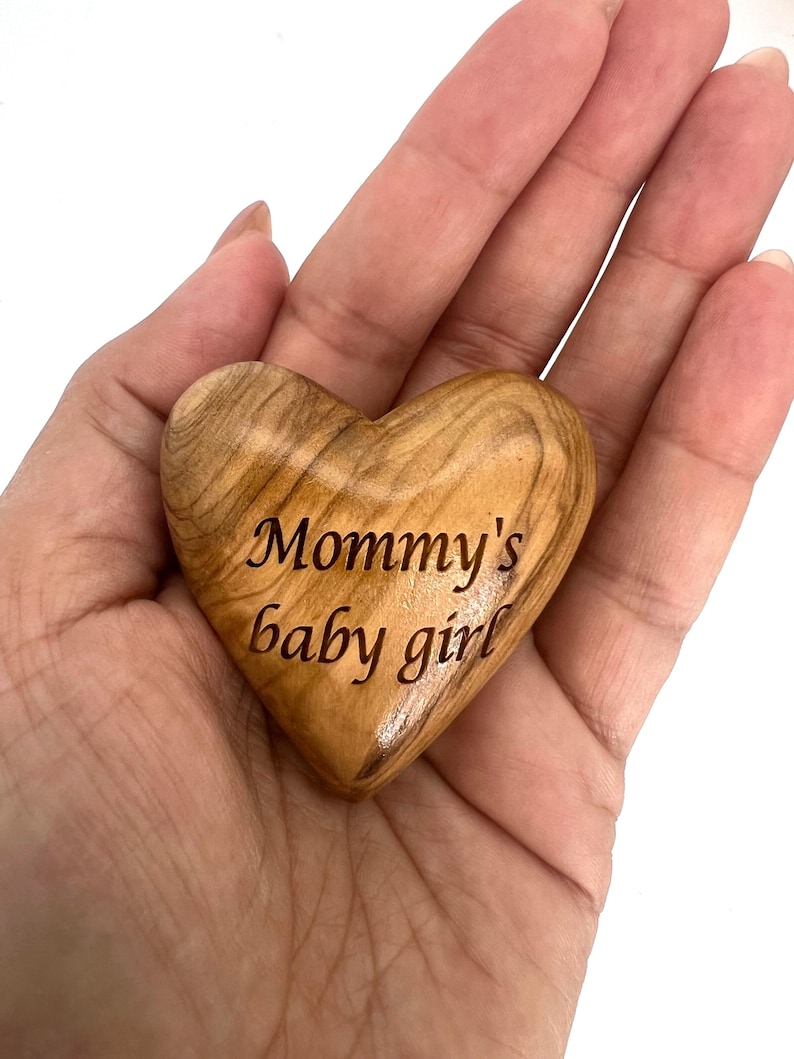 May include: A smooth, polished heart-shaped object carved from light brown wood with a visible natural grain pattern. The text "Mommy's baby girl" is engraved in a dark, cursive font across the center of the wooden heart surface.