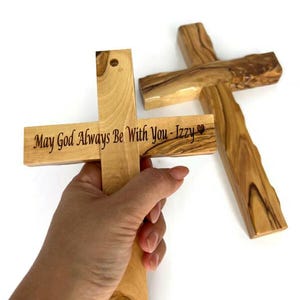 May include: Two wooden crosses, one held by a hand. The cross held has the inscription "May God Always Be With You - Izzy" in black lettering. The other cross is lying on a white surface. The crosses are made of light-colored wood.