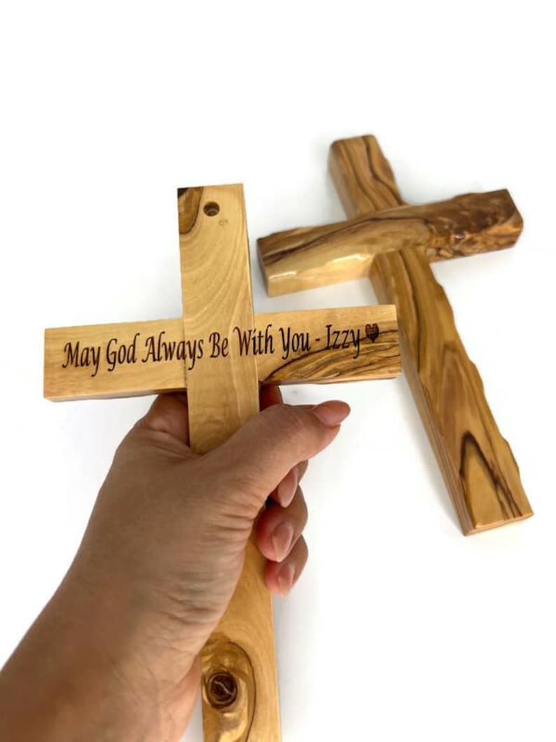 May include: Two wooden crosses, one held by a hand. The cross held has the inscription "May God Always Be With You - Izzy" in black lettering. The other cross is lying on a white surface. The crosses are made of light-colored wood.