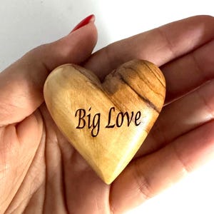May include: A smooth, polished wooden heart with a natural light brown grain and dark brown streaks. The words Big Love are engraved in a dark, cursive script on the front surface of the heart, which is shaped with a rounded top and pointed base.