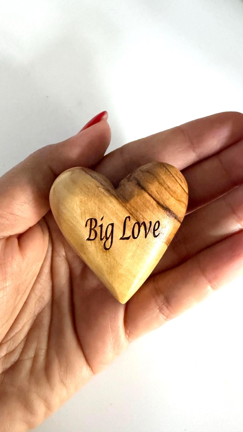 May include: A smooth, polished wooden heart with a natural light brown grain and dark brown streaks. The words Big Love are engraved in a dark, cursive script on the front surface of the heart, which is shaped with a rounded top and pointed base.
