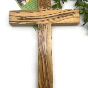May include: A wooden cross with a natural wood grain pattern. The cross has a vertical beam and a horizontal beam. A small card with text is attached to the cross. Green foliage is in the background.