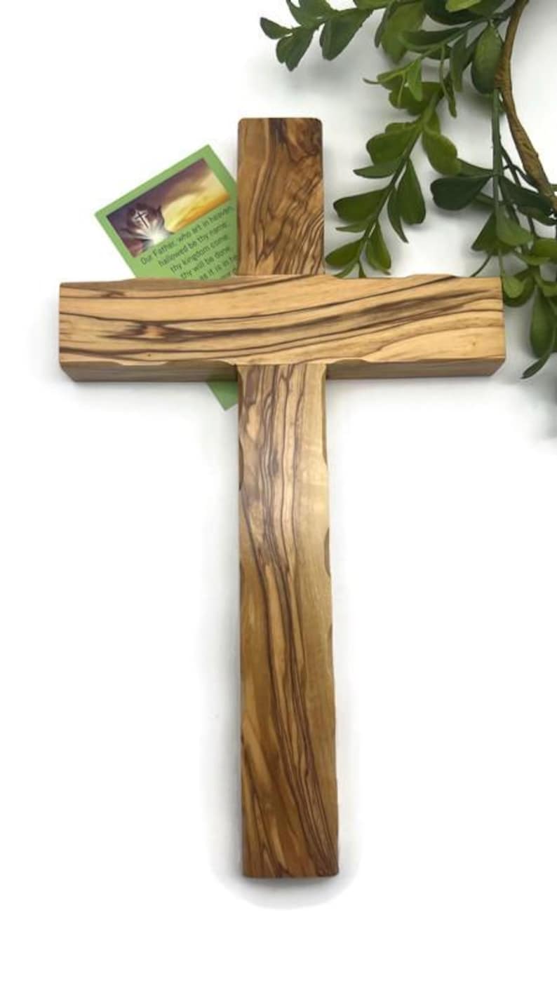 May include: A wooden cross with a natural wood grain pattern. The cross has a vertical beam and a horizontal beam. A small card with text is attached to the cross. Green foliage is in the background.