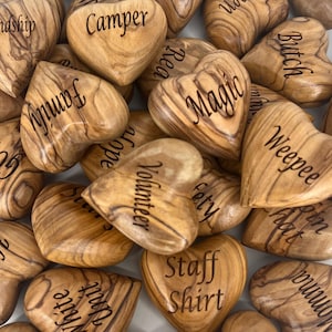 May include: A collection of polished olive wood hearts, each engraved with unique words including Craig, Yellow, Red, Fusco, Camper, Butch, Family, Magic, Weepee, Volunteer, Safety, Staff Shirt, White Unit, Inspire, and Ripley, showcasing natural grain.