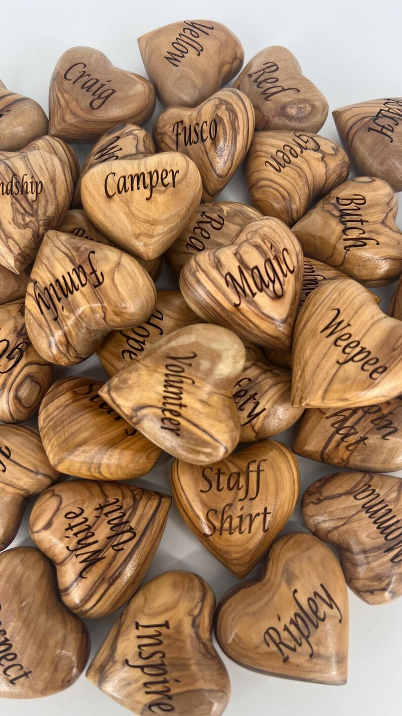 May include: A collection of polished olive wood hearts, each engraved with unique words including Craig, Yellow, Red, Fusco, Camper, Butch, Family, Magic, Weepee, Volunteer, Safety, Staff Shirt, White Unit, Inspire, and Ripley, showcasing natural grain.
