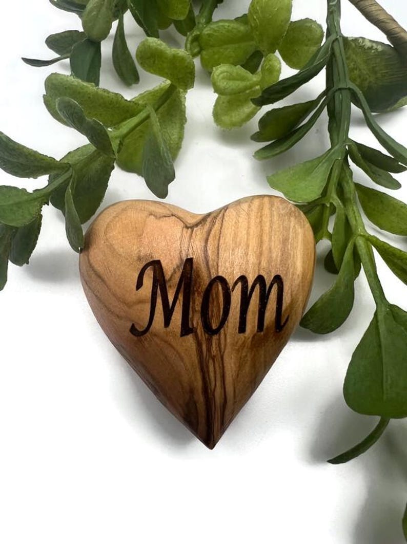 May include: A smooth, heart-shaped wooden object with a natural grain pattern and the word Mom engraved in a dark, cursive script on the front surface. The piece features light and dark brown wood tones with a polished finish and rounded edges.