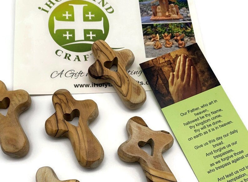 May include: Five small wooden crosses with a heart cutout in the center. The crosses are made of olive wood and have a natural wood finish. The crosses are arranged in a semi-circle on a white surface. The crosses are a religious symbol and are often used as a prayer aid.