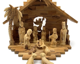 Può includere: Un presepe in legno con un presepe scolpito in dettaglio, figure di Maria, Giuseppe, Gesù bambino e i tre re magi, e due pecore. La scena si svolge in una stalla di legno con una palma e una stella sul tetto.