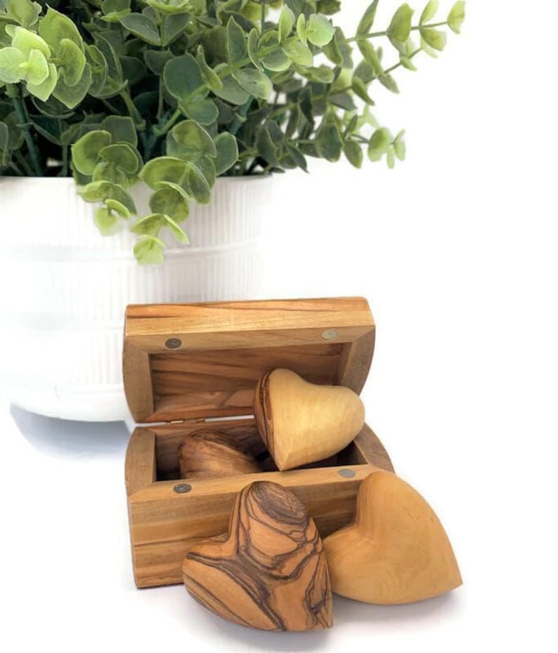May include: A small wooden chest with a hinged lid containing three smooth, heart-shaped wooden objects. The chest and hearts feature natural wood grain patterns in shades of light and dark brown, with visible magnetic closures on the chest frame.