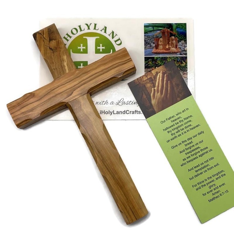 May include: A wooden cross with a natural wood grain finish. The cross is displayed with a bookmark featuring a prayer and the text "HOLYLAND" and "iHolyLandCrafts."