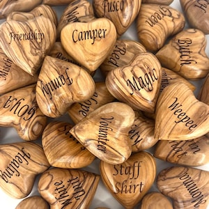 May include: A collection of smooth, heart-shaped wooden tokens crafted from olive wood, each featuring a unique grain pattern and a distinct word or name engraved in black text, including terms like Friendship, Camper, Magic, Family, and Respect.